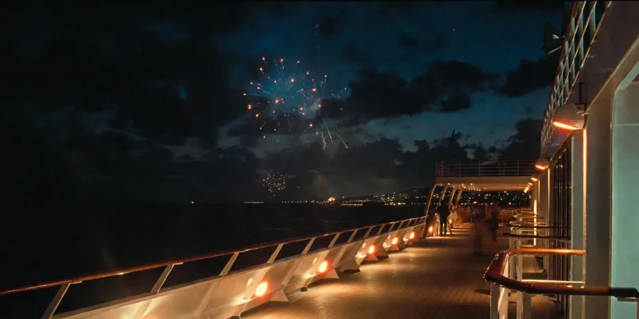 Night view on a cruise ship deck with warm lights and fireworks over the sea.
